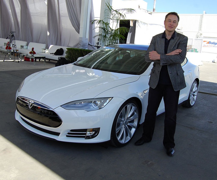 Elon Musk standing
beside a white Tesla car with his arms folded