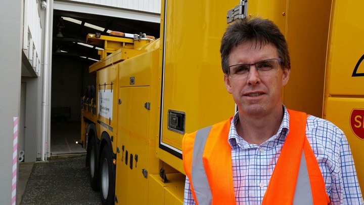 Mark Owen wearing
an orange safety vest, standing in front of a big yellow
vehicle