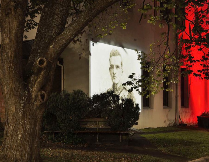 an old head and shoulders photograph of a soldier, projected onto the side of a building, seen through trees and bushes of a small plaza