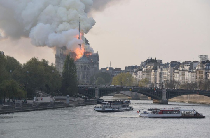 fire burning in
nortre dame cathedral, seem from the seine river with boats
and a bridge in the foreground. Flames are in the roof but
the spire is standing in this picture