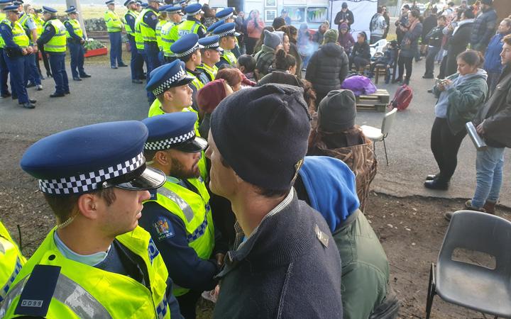 people facing off
with a line of police. More police and protestors stand
around behinds the lines. In the background some protestors
are on seats around a small fire