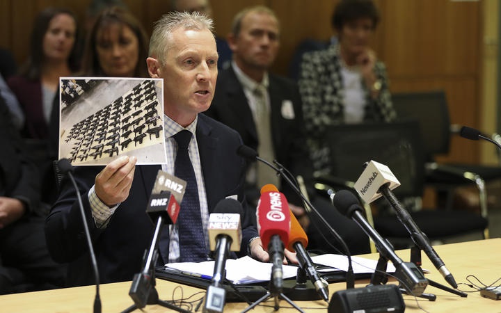Chris Cahill at a
desk with media microphones, holding up a photograph of a
lot of dangerous-looking guns