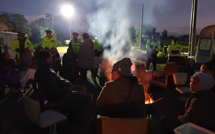 at night,protestors
sitting on seats arranged around a containedfire, in front
of a line of police