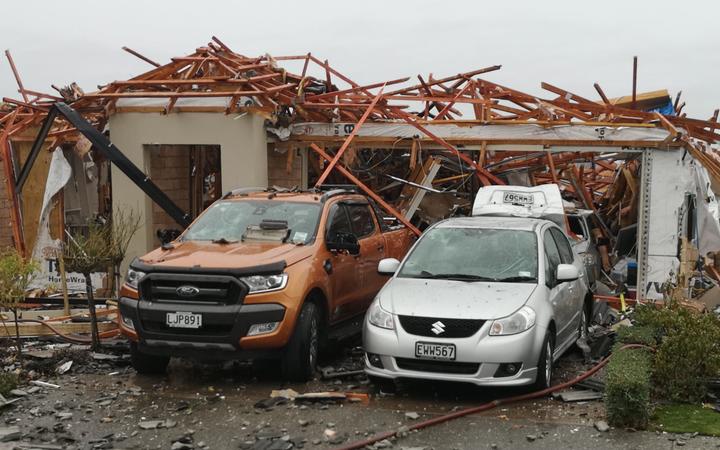 a house with no
roofing material and a collapsing roof framework, as well as
other damage. Debris is scattering all around. Two vehicles
are parked in front, one is a car with a boot the is lifted
and buckled.