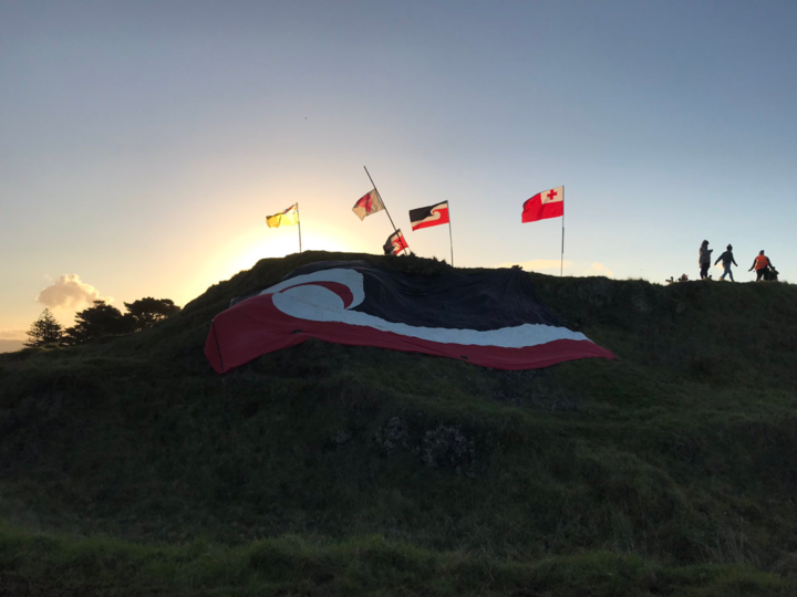 a very large tino
rangatiratanga flag draped on a hill, with other flags on
poles at the top and the sunset behind