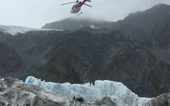 a helicopter hovers
with a line attached to a large package on the glacier, with
a person touching the bundle's side