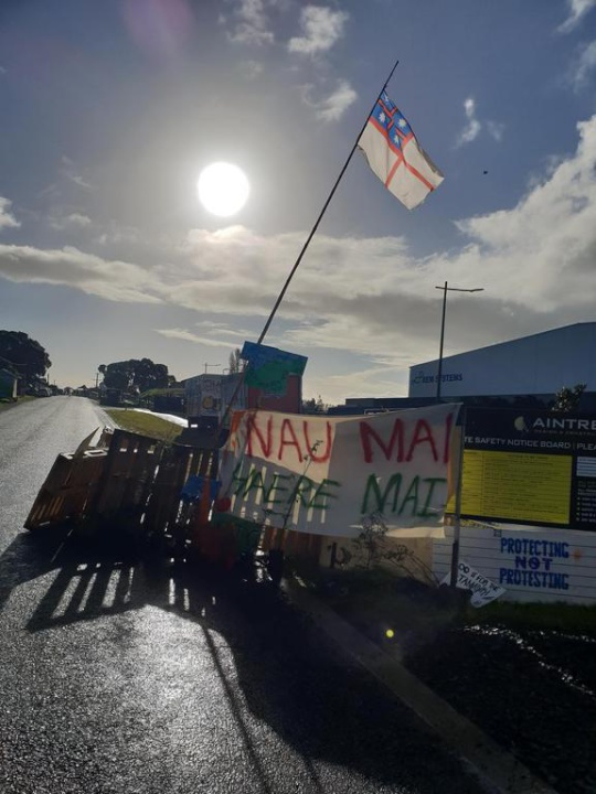 an access road
partly blocked by a shipping palette fence, with a united
tribes flag on a pole and a large banner reading Nau Mai
Haere Mai