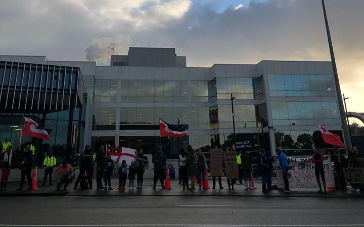 protestors lined up
bside the road with māori flags, placards, and a banner