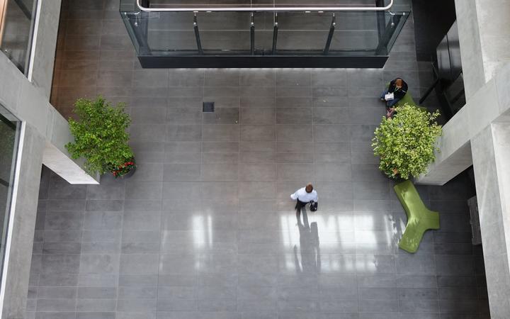 an atrium viewed
from above, with smooth stone flooring, two trees in pots,
and square concrete columns around it. One person is walking
through with a briefcase and another is on the seating