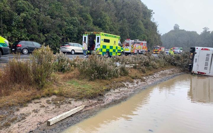 a bus on its side
in a shallow river bed, on the road emergency vehicles and
stopped cars